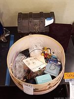 Round floral cardboard box filled with assorted jewelry; wooden trunk and velvet case visible in background