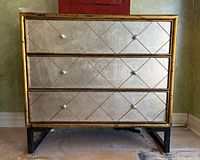 Front view of three-drawer dresser showing silver drawers with diamond pattern and gold frame, resting on black metal legs.
