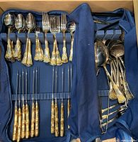 Overview of multiple forks, spoons, and serving utensils with gold-toned ornate handles organized in a blue fabric flatware case.