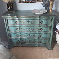 Front view of the turquoise distressed 4-drawer dresser with gold ornamental details and ring pull handles.