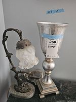 Photo showing Victorian cherub swing lamp with frosted flower-shaped shade next to chalice-shaped silver-toned lamp.