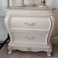 Front view of the cream Bombay style nightstand showing detailed carvings, curved drawers with ornate handles and marble-like top with decorative items.