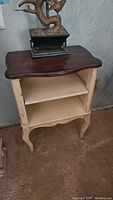 Side view of wood side table shelf showing dark wood shaped top and cream painted distressed frame with two shelves and cabriole legs