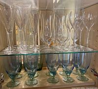 Shelf displaying clear crystal champagne flutes, wine glasses, and blue-tinted water glasses with thick stems and bases.