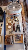 Overhead view of ultrasonic cleaner on tray with assortment of wrist and pocket watches, wooden watch box, straps and gemstone bag