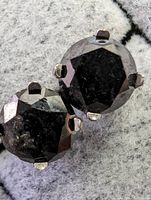 Close-up photo of one black diamond earring shows round faceted diamond held by four white gold prongs.