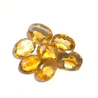 Seven faceted oval citrine gemstones in a golden yellow to orange hue arranged together on a white background for clarity and color assessment.