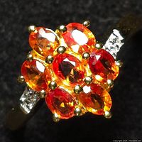 Close-up front view showing nine faceted oval orange sapphires arranged in a cluster with gold prongs on silver band with small round diamond accents on band sides.