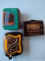 Photo showing three Sony Walkman cassette players on a flat surface: green Sports model, black FM/AM digital tuner model, and black/yellow Sports model.