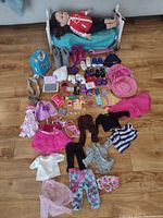 Full overview of American Girl doll laying on a metal-frame bed with multiple clothing, shoes, and accessories spread in front including food items, books, and small toys.