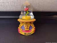 Front view of the Belle snowglobe showing a red rose under a glass dome on a decorative yellow and purple base labeled 'Belle'.