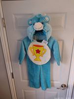 Blue Care Bear costume hanging on a door, showing the front with hood up, trophy emblem on belly.