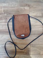 Brown leather front flap purse with black trim and long strap, laid flat on wooden floor.