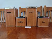 Front view of four kitchen canisters labeled flour, sugar, coffee, and tea with wood-look finish and chrome lids.