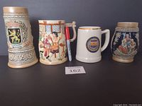 Front view of four ceramic steins and mug on table with pen for scale, shows details of relief work and emblem on mug.