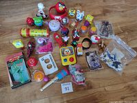 Full overview of the lot showing various toys on a wooden floor including a metal kaleidoscope, boxed figure, plush Angry Birds, multiple plastic toys and figures, and some items in packaging or plastic bags.