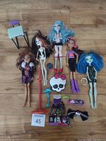 Photo showing five Monster High dolls and assorted accessories on a wooden floor including a desk, shoes, and themed items.