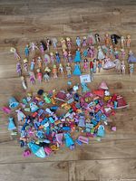 Wide view of 44 dolls laid in rows with a large pile of small clothing and accessory items below them on a wood floor