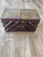 Front view of closed wooden sewing box showing floral carvings on lid and accordion style side arms.