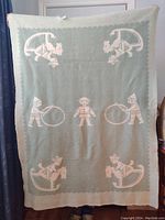Front side of vintage baby wool blanket held up for display showing light green background with cream-colored toy soldier, rocking horse riders, rabbits, and children holding hoops