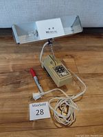 Full view of vintage headboard lamp showing rectangular white lamp housing with two sockets on beige base with floral graphic and attached cord with plug next to a marker for scale.