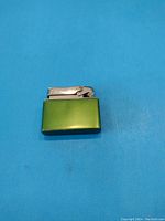 Front view of the green metal vintage lighter with silver ignition cap against a blue background.