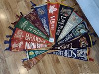 Full view of 13 vintage travel pennant banners arranged on floor, mostly felt, different colors and cities, including one small leather banner.