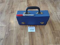 Blue vintage vinyl cassette holder with a handle and two gold-tone clasps on front sitting on a wooden floor with a red Sharpie marker placed on top.