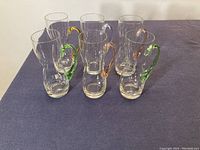 Six clear tall drinking glasses with colored handles in green, yellow, and amber, arranged in a group on a dark surface.