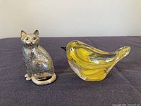Silver cat figurine sitting, showing engraved fur texture, amber eyes, and curled tail, with yellow glass bird beside