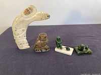 Four carved items on a dark cloth: large beige human-animal figure, brown owl carving, two small green stone figures.