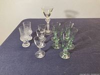Overview of eleven assorted Scandinavian cordial glasses, including clear textured, clear smooth, and green glasses with bulbous stem details arranged on a dark surface.
