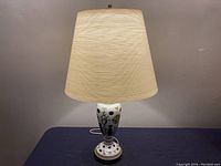 Full view of the vintage porcelain lamp with a white fabric shade lit on a table with dark cloth background.