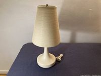 Full view of lamp showing beige fabric shade, white matte base, and attached cord