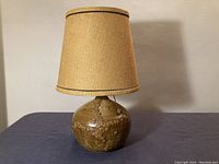 Full view of vintage pottery table lamp with beige fabric shade sitting on a blue cloth surface.