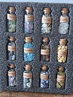 Front view of 12 glass bottles containing various labeled gemstones, arranged in foam holder.