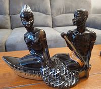 Photo showing black glazed ceramic figurine depicting two stylized figures in a canoe with detailed texture, with the figurine placed on a wooden surface and photographed in natural indoor lighting.