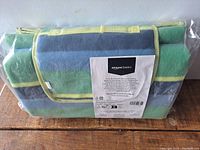 Folded picnic blanket in plastic packaging showing Amazon Basics label with product info and care instructions.