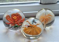 Photo showing a set of three clear glass paperweights each with a detailed internal orange floral or insect design, displayed on a white surface by a window.
