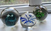Photo showing three glass paperweights on a white surface in front of a window, illustrating the internal blue and green designs of each sphere.