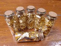 Six small glass vials with cork stoppers filled with gold leaf flakes sitting on a wooden surface.