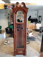 Full view of the Herschede tall wooden hall clock with glass door and curved top, standing on a garage floor surrounded by storage boxes and furniture.