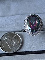 Silver ring size 7 with an oval mystic topaz surrounded by a halo of clear stones placed beside a Canadian dime for size reference, showing the ring on a fabric background.