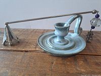 Light blue-gray glazed pottery candle stick with handle placed on a wooden surface. Attached is a metal candle snuffer with a beaded charm.