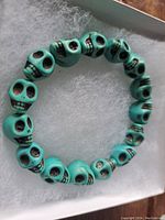 Top-down view of the turquoise colored stone skull carved stretch bracelet laid on white padding inside a box.