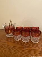 Set including six short cocktail glasses with red and gold band and clear thick bases, arranged with a clear glass ice bucket and clear ice tongs on a wooden surface.