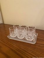 Set of six crystal shot glasses arranged on a rectangular crystal tray on a wooden surface against a beige wall.