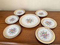 Photo showing six round plates surrounding one large matching serving platter, all white porcelain with intricate gold borders and multicolored floral center design.