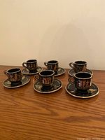 Set of six black espresso cups with gold Grecian figures and matching saucers on wood surface