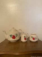 Photo showing front view of the three-piece ceramic tea set on wooden surface against plain wall. Includes teapot, milk creamer, and sugar container, all white with red floral decoration.